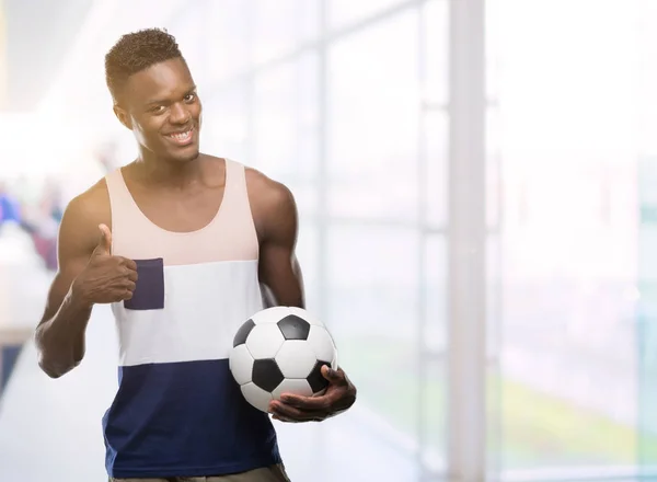 Young african american man holding soccer football ball happy with big smile doing ok sign, thumb up with fingers, excellent sign