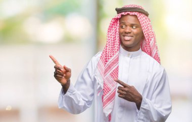 Gülümseyen ve iki elleri ve parmakları ile tarafına işaret kamera bakarak izole arka plan üzerinde geleneksel keffiyeh giyen genç Arapça Afrika adam.