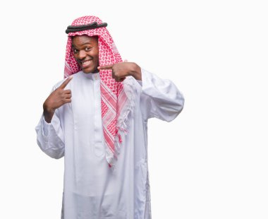 Geleneksel keffiyeh kendine güvenen gösterilen ve işaret parmakları diş ve ağız ile gülümseyen izole arka plan üzerinde giyen genç Arapça Afrika adam. Sağlık kavramı.