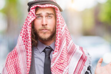 Genç yakışıklı Arap adam uzun saçlı keffiyeh izole arka plan clueless ve şaşkın bir ifadeyle kollar ve eller kaldırdı üzerine giyiyor. Şüphe kavramı.