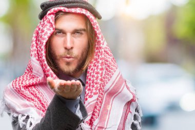 Genç yakışıklı Arap adam uzun saçlı bir öpücük el ile güzel ve seksi olmak hava üfleme kamera bakarak izole arka plan üzerinde keffiyeh giyiyor. Aşk ifade.