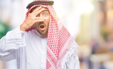 Mahcup ifadeyle parmaklarımın arasından seyir keffiyeh yüz ve gözler el ile kapsayan şok bakmak izole arka plan üzerinde giyen genç yakışıklı adam.