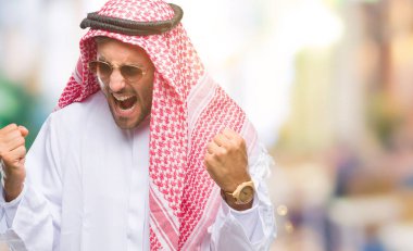 Gülümseyen ve başarı için çığlık keffiyeh izole arka plan çok mutlu ve heyecanlı kaldırdı, silah ile kazanan jest yapıyor üzerinde giyen genç yakışıklı adam. Kutlama kavramı.