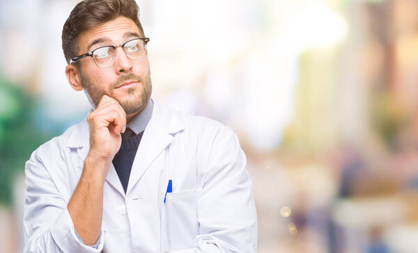 Young handsome man wearing doctor, scientis coat over isolated background with hand on chin thinking about question, pensive expression. Smiling with thoughtful face. Doubt concept.