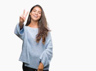 Mutlu yüz zafer işareti yaparken kameraya göz kırpıyor ile gülümseyen izole arka plan üzerinde kış kazak giyen genç güzel Arap kadın. İki numaralı.