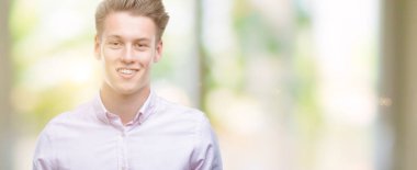 Genç yakışıklı sarışın adam ayakta ve diş gösterilen kendine güvenen bir gülümsemeyle gülümseyerek mutlu bir yüz ile