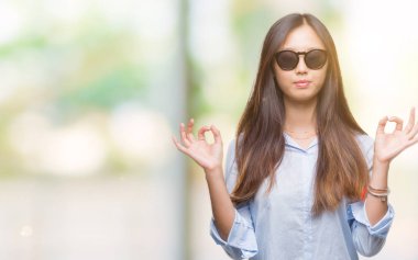 Genç Asyalı kadın giyiyor güneş gözlüğü izole arka plan üzerinde dinlenmek ve gülümseyen gözlerle kapalı parmak jestle meditasyon yapıyor. Yoga kavramı.