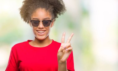 Mutlu yüz zafer işareti yaparken kameraya göz kırpıyor ile gülümseyen izole arka plan üzerinde güneş gözlüğü takmış genç afro Amerikalı kadın. İki numaralı.