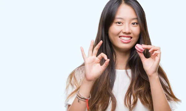 優秀なシンボルの指で サインをして孤立の背景にピンクのマカロン甘いを食べる若いアジア女性 — ストック写真
