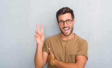 Mutlu yüz zafer işareti yaparken kameraya göz kırpıyor ile gülümseyen gözlüklü gri grunge duvarın üzerinden yakışıklı genç adam. İki numaralı.