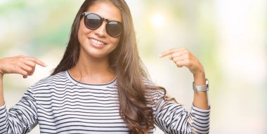 Genç güzel Arap kadın yüzünde gülümseme ile kendinden emin görünüyorsun, kendini gururlu ve mutlu parmakları ile işaret izole arka plan üzerinde güneş gözlüğü takıyor.