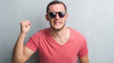 Güneş gözlüğü rahatsız ve hayal kırıklığına uğramış bağırarak öfkeyle, çılgın ve yükseltilmiş el, öfke kavramı ile diye bağıran giyen gri grunge duvarın üzerinden beyaz delikanlı