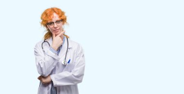 Genç Kızıl saçlı kadın giyen Doktor emin çapraz kolları olan gülümseme ile kameraya bakarak üniforma ve üzerinde yükseltilmiş çene el. Pozitif düşünce.