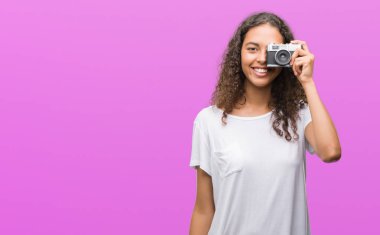 Genç İspanyol kadın ayakta ve diş gösterilen kendine güvenen bir gülümsemeyle gülümseyerek mutlu bir yüz ile Vintage kamera ile fotoğraf çekmek