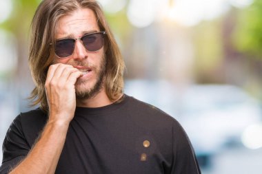 Stresli ve gergin ile izole arka plan üzerinde güneş gözlüğü takmış uzun saçlı yakışıklı delikanlı tırnak ısırma dudaklarından eller. Anksiyete sorunu.