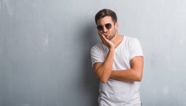 Yakışıklı genç gri grunge üzerinde duvar düşünme seyir yorgun ve çapraz kolları olan depresyon sorunları ile sıkılmış giyen güneş gözlüğü.