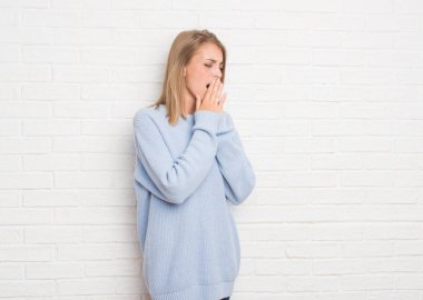 Güzel genç kadın beyaz tuğla duvarın üzerinden el ile ağız kapsayan yorgun esneme sıkılmış. Restless ve uyku hali.