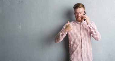 Genç Kızıl saçlı adam telefonda büyük mutlu ile konuşurken gri grunge duvarın üzerinden gülümseme ok işareti, başparmak ile parmak, mükemmel işareti yapıyor
