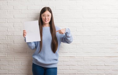 Genç Çinli kadın kendisine sürpriz yüz işaret parmağı boş kağıt tutan tuğla duvarın üzerinden