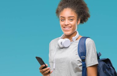 Sırt çantası izole arka plan üzerinde duran ve diş gösterilen kendine güvenen bir gülümsemeyle gülümseyerek mutlu bir yüz tutan genç güzel afro Amerikan öğrenci kadın