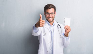 Yakışıklı genç doktor adam gri grunge boş defter ok işareti yapıyor büyük gülümseme ile mutlu tutarak duvar, parmaklarıyla, mükemmel işaret parmak