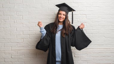 Duygu tezahürat lisansüstü Tekdüzen gurur çığlık ve zafer ve başarı çok heyecanlı, kutluyor beyaz tuğla duvar üzerinde duran genç esmer kadın