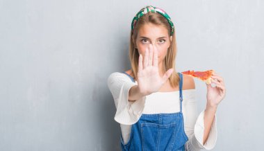 Grunge gri duvar açık eliyle dur işareti ile ciddi ve kendine güvenen ifade, savunma jest yapıyor pepperoni pizza dilimi yemek güzel genç kadın