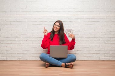 Yerdeki beyaz tuğla üzerinde oturan genç esmer kadın ödeme yapan holding dolar bir fikirle sürpriz duvar veya gülen yüz, bir numara ile işaret parmağı soru