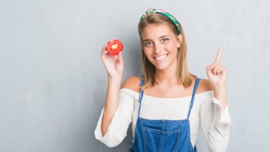 Grunge güzel genç kadın taze kırmızı domates dilim bir fikirle sürpriz yemek duvar gri veya gülen yüz, bir numara ile işaret parmağı soru