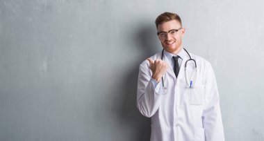 Genç redhead Doktor adam işaret ve başparmak kadar mutlu yüz gülümseyen tarafı ile gösterilen gri grunge duvarın üzerinden