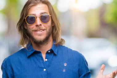 Genç yakışıklı adam uzun saçlı izole arka plan clueless ve şaşkın bir ifadeyle kollar ve eller kaldırdı üzerinde güneş gözlüğü takıyor. Şüphe kavramı.