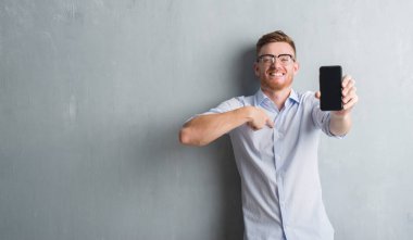 Genç Kızıl saçlı adam gri grunge duvar gösteren boş ekran kendisi için sürpriz yüz işaret parmağı ile Smartphone