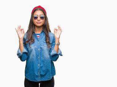 Genç güzel Arap kadın giyiyor güneş gözlüğü izole arka plan üzerinde dinlenmek ve gülümseyen gözlerle kapalı parmak jestle meditasyon yapıyor. Yoga kavramı.