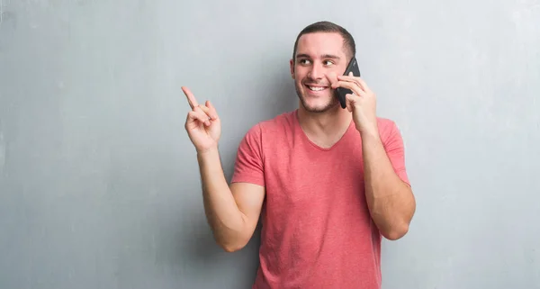 Молодой Кавказский Человек Серой Гранж Стеной Разговаривающий Телефону Доволен Указывая — стоковое фото