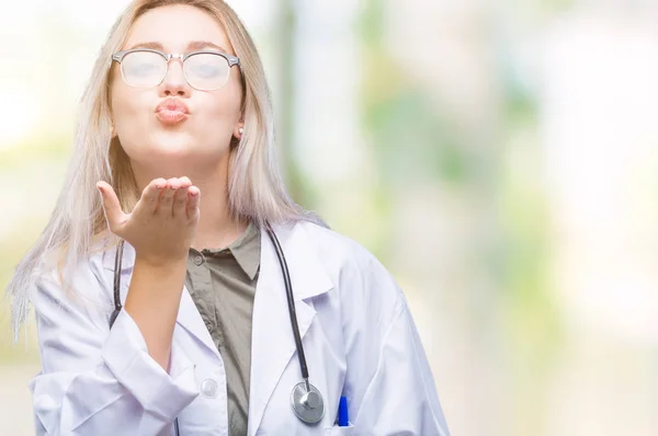 Young blonde doctor woman over isolated background looking at the camera blowing a kiss with hand on air being lovely and sexy. Love expression.