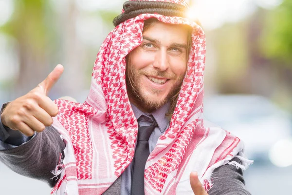 年轻英俊的阿拉伯男子与长发穿着 Keffiyeh 在孤立的背景下批准做积极的手势与手 竖起大拇指微笑 并为成功而高兴 看着相机 胜利者的手势 — 图库照片