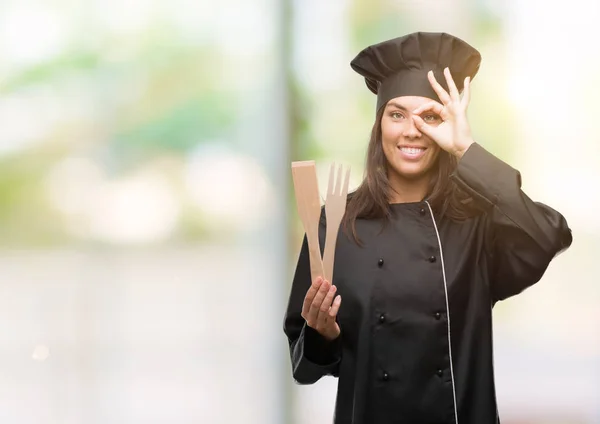Young hispanic cook woman wearing chef uniform with happy face smiling doing ok sign with hand on eye looking through fingers