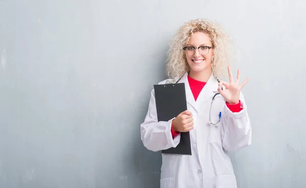 年轻的金发医生女人超过垃圾灰色墙壁持有剪贴板做 标志与手指 优秀的符号 — 图库照片
