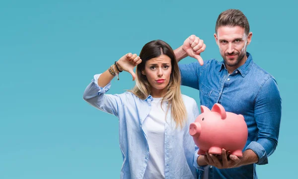 Young couple in love holding piggy bank over isolated background with angry face, negative sign showing dislike with thumbs down, rejection concept