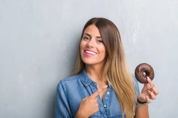 チョコレート ドーナツの手と指で指している非常に幸せを食べる灰色グランジ壁を越えて若い成人女性 — ストック写真