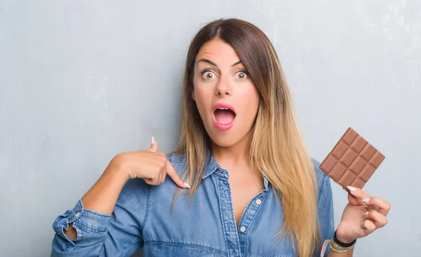 灰色のグランジ壁自分自身に驚き顔ポインティング指でバー チョコレートを食べることで若い成人女性 — ストック写真
