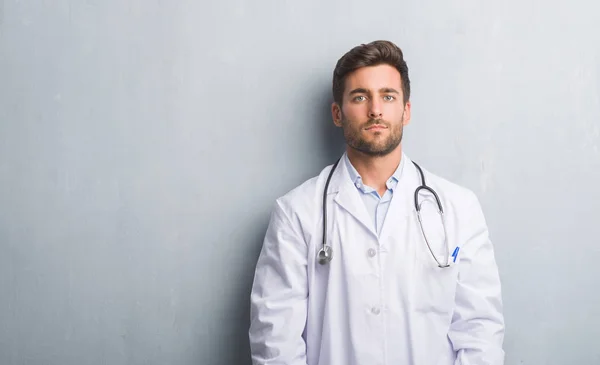 Handsome young doctor man over grey grunge wall with serious expression on face. Simple and natural looking at the camera.