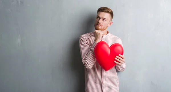 Young Redhead Man Grey Grunge Wall Holding Red Heart Serious — Stock Photo, Image