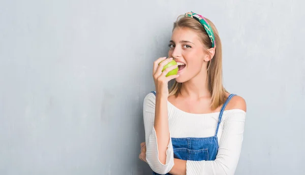 スマートの顔深刻な思考に自信を持って表現と青リンゴを食べてグランジ灰色の壁を越えて美しい若い女性 — ストック写真