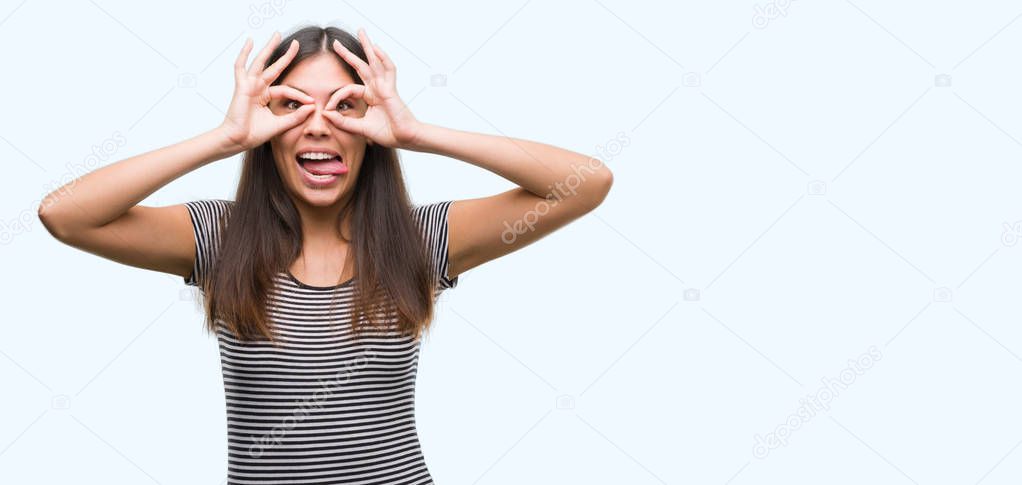 Young beautiful hispanic woman doing ok gesture like binoculars sticking tongue out, eyes looking through fingers. Crazy expression.