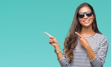 Genç güzel Arap kadın gülümseyerek ve iki elleri ve parmakları ile tarafına işaret kamera bakarak izole arka plan üzerinde güneş gözlüğü takıyor.