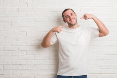 Kendinden emin gösterilen ve işaret parmakları diş ve ağız ile gülümseyen beyaz tuğla duvarın üzerinden ayakta beyaz delikanlı. Sağlık kavramı.