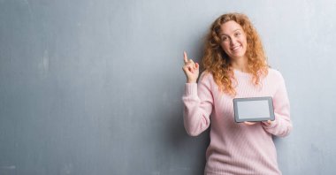 El ve parmak ile tarafına işaret çok mutlu tablet kullanarak gri grunge duvar genç Kızıl saçlı kadın