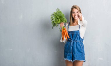 Taze havuç kamera ve size parmak ile işaret tutan grunge gri duvarın üzerinden güzel genç kadın, el işareti, pozitif ve kendinden emin bir jest ön