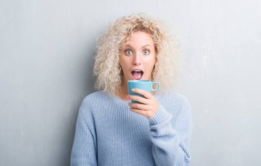 Grunge gri duvar driking bir fincan kahve bir sürpriz yüz, korkuyor ve heyecanlı korku ifadesi ile şok korkmuş genç sarışın kadın
